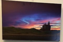 A landscape featuring distant mountains and a barn, both darkened by the encroach of the evening. The sky in contrast is a blaze in brilliant blues and purples with a faint yellow peeking out from behind the mountains as the sun sets.