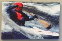 A kayaker wearing blue gear, a red life vest and a red helmet is captured mid paddle, the implement blurred in motion, surrounded by white rapids.  