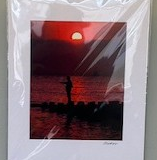 The sun sets beneath the dark silhouette of a mountain range, lighting up the sky in crimson which is reflected upon the surface of a nearby shore.  Someone stands on some rocks jutting out into the water, shrouded in darkness.