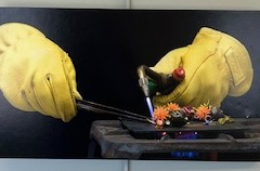 A closeup on the hands of a glassworker. They are wearing thick yellow heat resistant gloves with the left holding tongs and the right holding a blow torch. They manipulate a small glass ornament of plant on a table. 