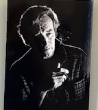 An elderly man holds a cigarette. Captured in greyscale, a harsh lighting effect illuminates one side of the man while shrouding the other in darkness.