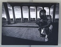 A shirtless man kneels. The bricks under his feet are arranged in a zig zag pattern, extending to a stone fence marked by tall Roman columns. The sky is visible around the construct. Captured in greyscale. 