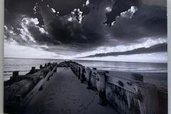 A narrow strip of sandy land, surrounded by sturdy rocks juts out into the ocean. An old but thick and sturdy fence lines the perimeter. Dark clouds loom overhead in a pure white sky. Captured in greyscale.