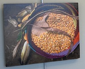 Several ears of corn, some shucked and some still bearing their green husks, lay on the ground around a maroon bowl featuring a swirling pattern of yellows, blues, oranges, and purples. The bowl is filled with corn kernels, and topped with a corn tassel laying upon the kernels across the center of the bowl.